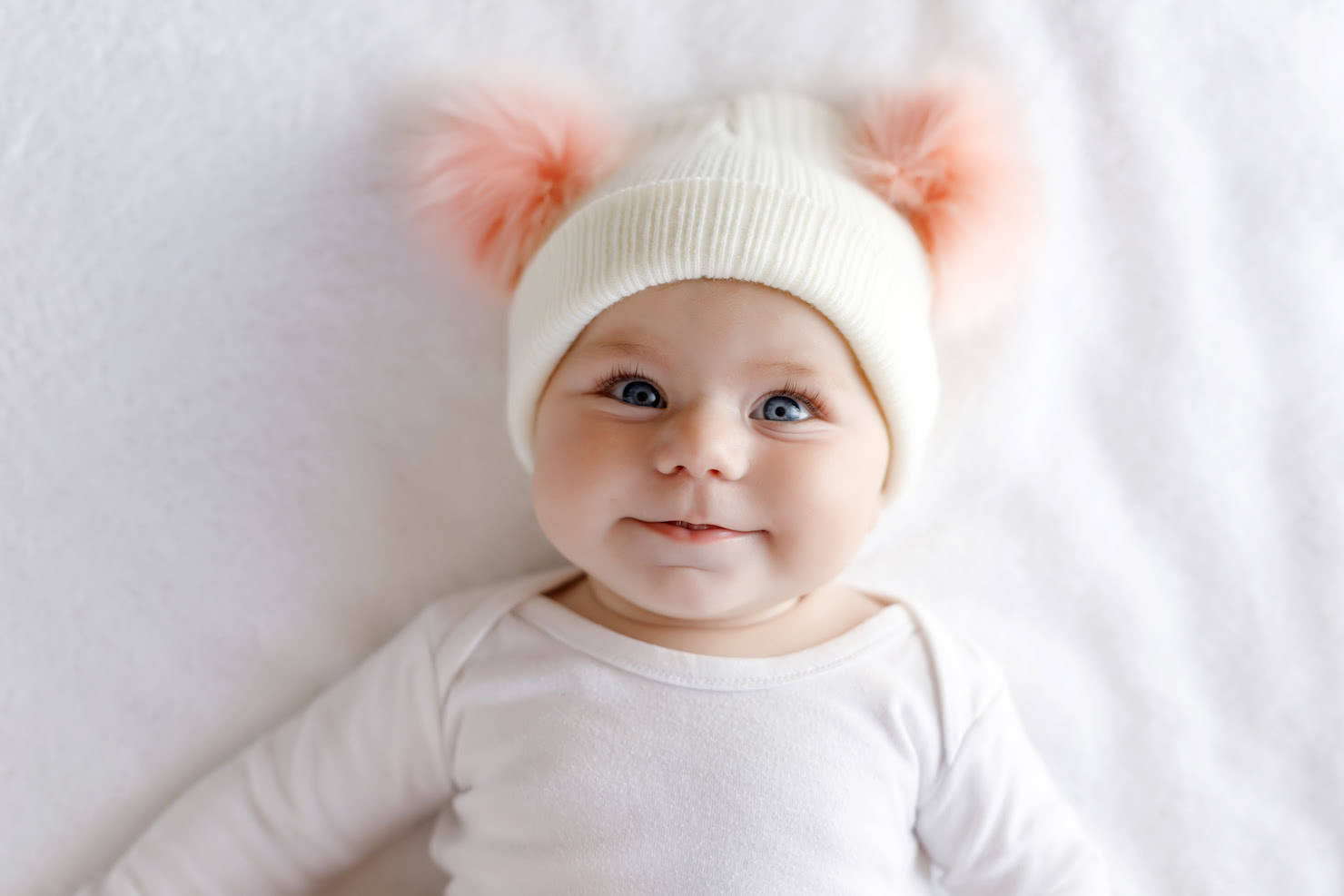 High angle shot of a baby girl laying on white blanket wearing a cream colored beanie with pink poms