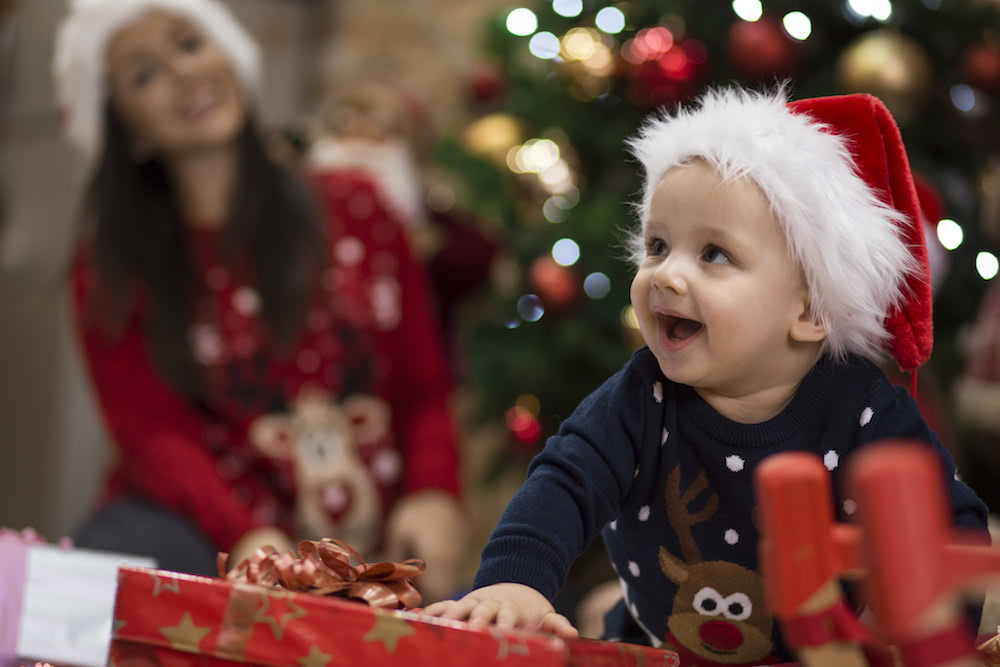 Baby wearing a red santa claus hat playing with a christmas present while the joyful mother watches