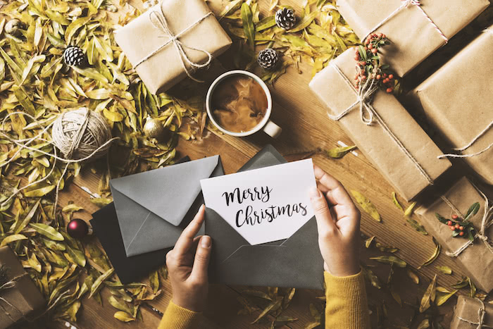 Woman sending a merry christmas card in a gray envelope featuring four brown paper wrapped gifts