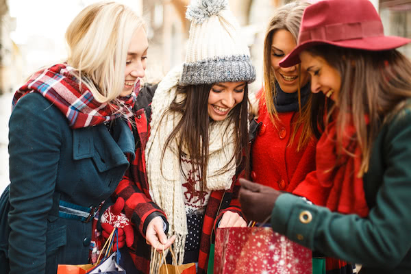 Four happy and curious women dressed in winter outfits and hats looking inside a red shopping bag with festive decorations