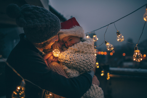 Young girl wearing a santa claus hat sitting on the lap of their mother while they admire hanging light bulbs outdoors