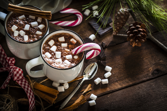 Two white mugs of hot chocolate with marshmallows featuring two red and white candy canes and two pinecones 