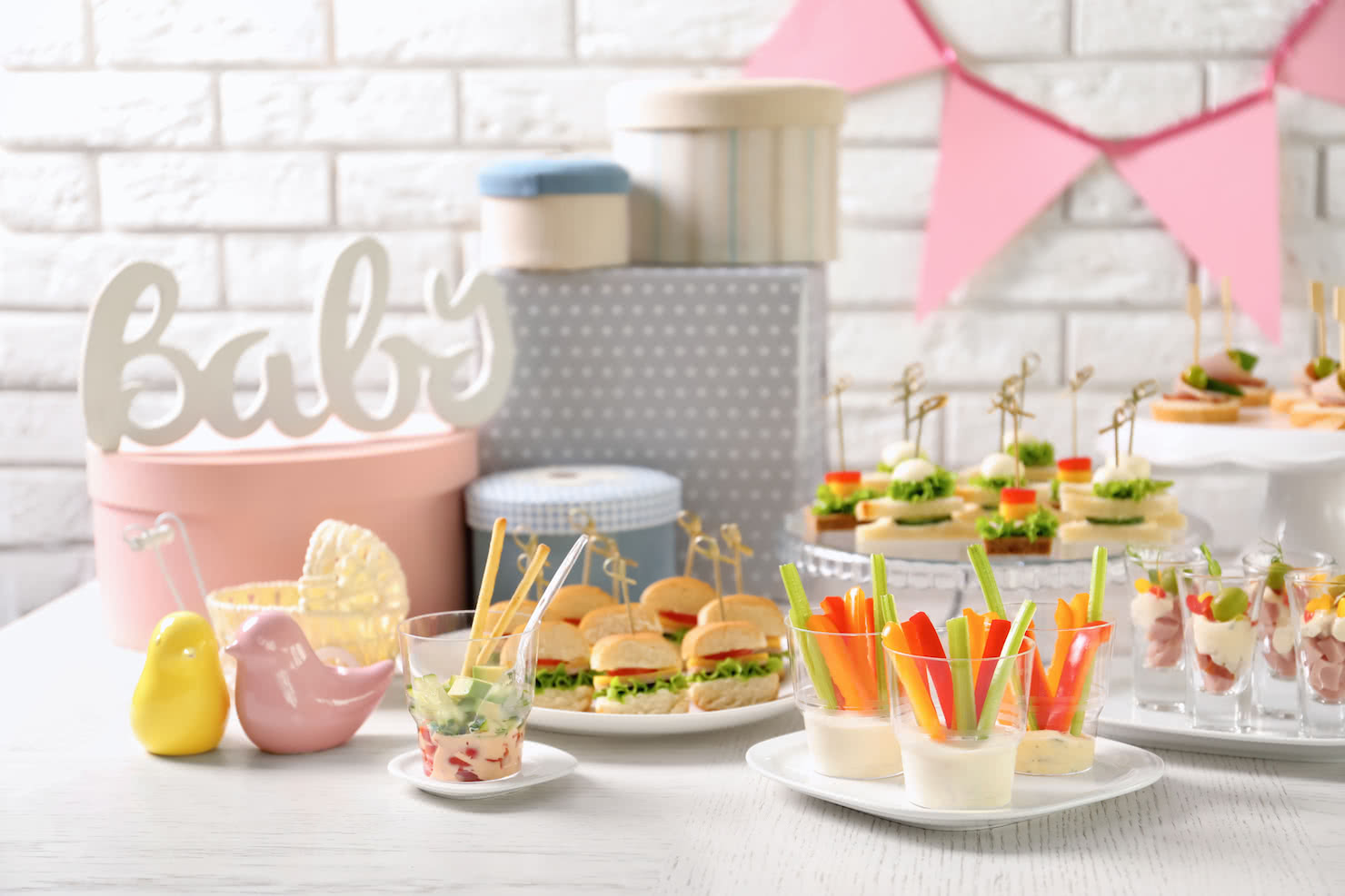 Table featuring sandwiches, finger foods, and baby shower gifts with a pink bunting banner hanging on a white brick wall