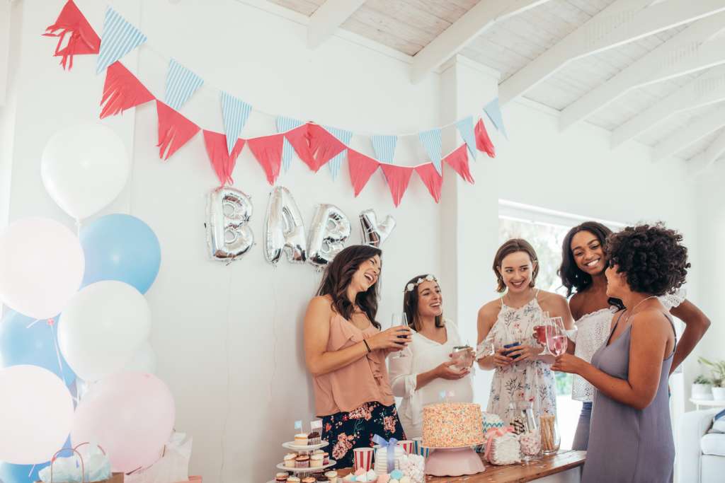 Five women celebrating a baby shower at home featuring silver balloons that spell baby and colorful pink and blue banners